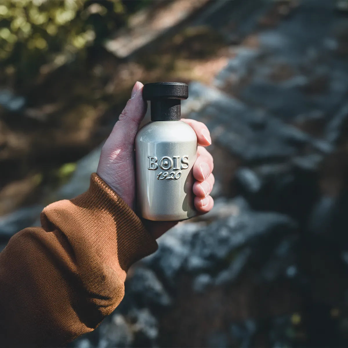 Hand holding a Bois 1920 perfume bottle against a blurred natural background