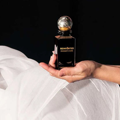 Hand holding a black perfume bottle labeled 'Reinvented Enigmatic Aura' against a dark background.