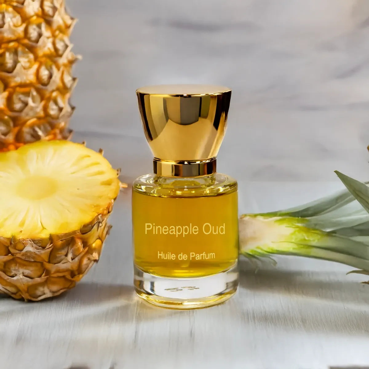 Pineapple Oud fragrance bottle placed beside fresh pineapple halves on a marble surface