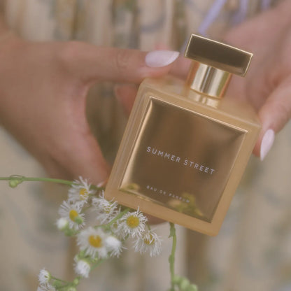 Gold 'Summer Street' perfume bottle held by a hand with flowers in the foreground